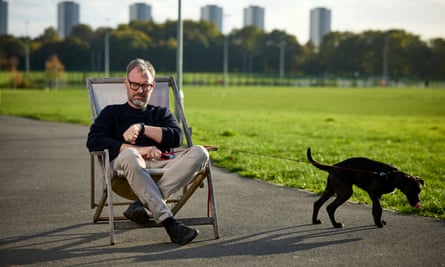 Tim Dowling, on a deckchair in a park, looks at his watch, while his dog, on a lead, sniffs at the edge of the grass