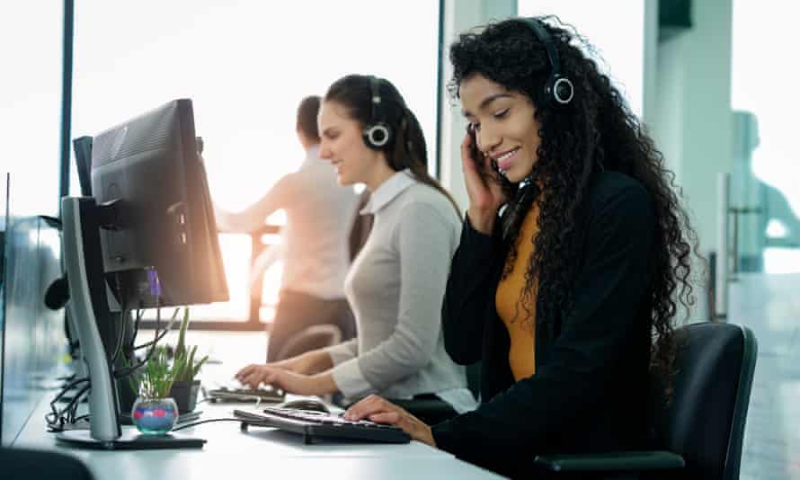 Women sitting in an office, doing call centre work, smiling in front of the computer, one very focused on her work, the other one smiling at the camera