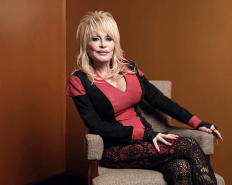 Dolly Parton sits in an armchair, wearing a red and black top and lace trousers