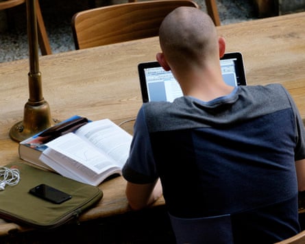 Student photographed from behind, sitting at a table on a laptop with an open book propper up next to them