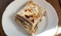Square slice of five-layer lasagne on a slightly scalloped plate in front of a porcelain tray of lasagne, both resting on round woven trivets on a wooden table