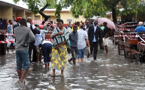 People walk through ankle-deep flood waters next to a polling station, with wooden benches piled up to one side and cordoned off