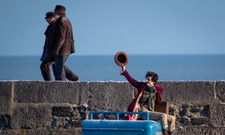Timothée Chalamet in costume as Willy Wonka waves with his hat while two men walk by on the top of an old stone harbour wall, with the sea and blue sky in the background
