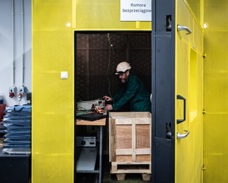 A man wearing protective gear works at a cluttered desk inside a bright yellow enclosed chamber, adjusting electronic equipment and cables.