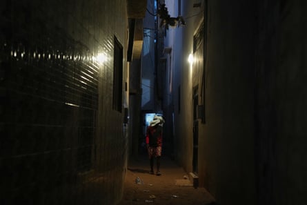 A man with features concealed behind a scarf stands in the shadows in a dark alley.