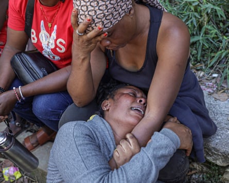 A woman holds her head in her hand as her other arm is gripped by a weeping woman whose head is in her lap