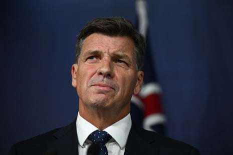 A man with dark hair in a dark suit stands before a flag.