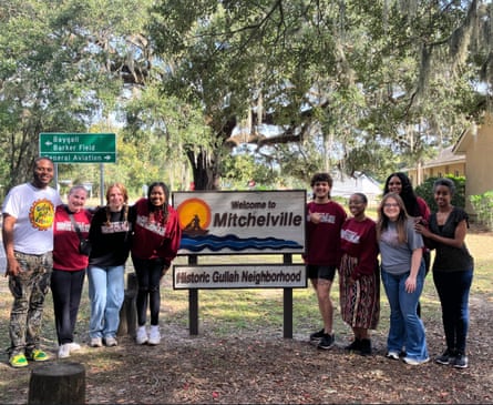 Nine people, mostly Black, stand to both sides of a park sign that says ‘Welcome to Mitchelville, Historic Gullah Neighborhood’, in shade among trees.