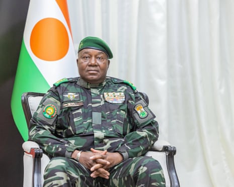 An older African man in camouflage fatigues and a beret sits by a green and orange flag