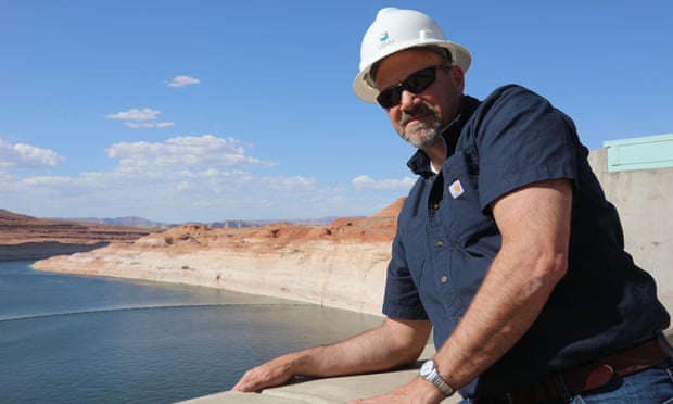 Man wearing a blue shirt and white helmet. In the background, a river sneaks around a canyon