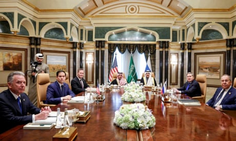 Οι άνδρες κάθονται γύρω από ένα μεγάλο, μεγαλοπρεπές τραπέζι, με έναν χειριστή κάμερας να στέκεται πίσω από τον Ρούμπιο