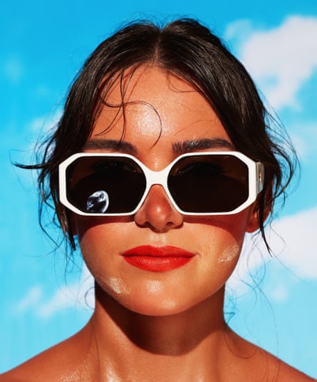 Headshot of a woman wearing white-framed sunglasses, with a smear of subncream on them, and sand on her face