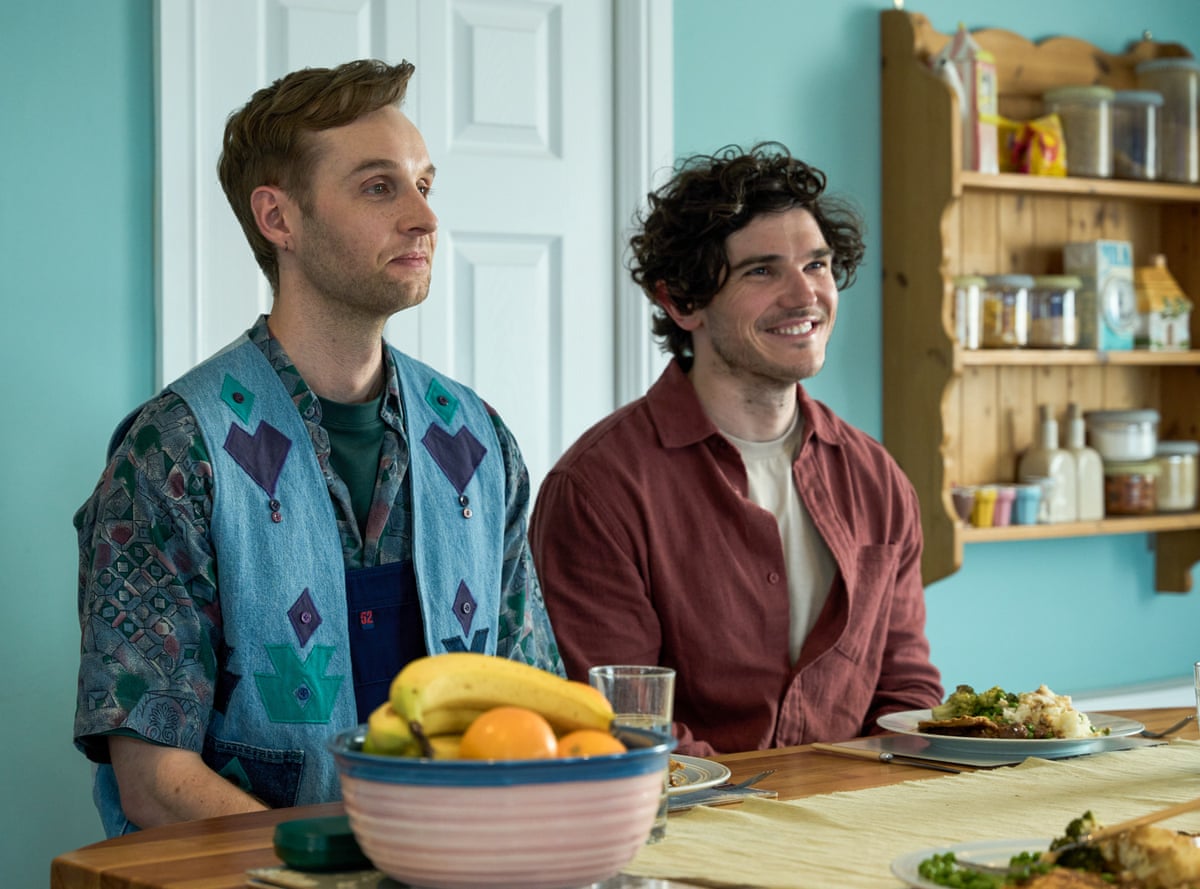 Two male actors from Lost Boys & Fairies sit at a kitchen table with a door and shelving behind them and fruit in a bowl and other food on the table