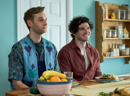 Two male actors from Lost Boys & Fairies sit at a kitchen table with a door and shelving behind them and fruit in a bowl and other food on the table