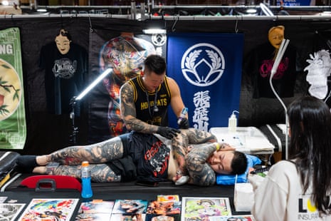 A person with tattooed arms and legs lies on their side while a tattooist tattoos them