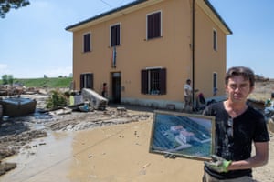 A person holding a framed picture stands among debris in front of a building