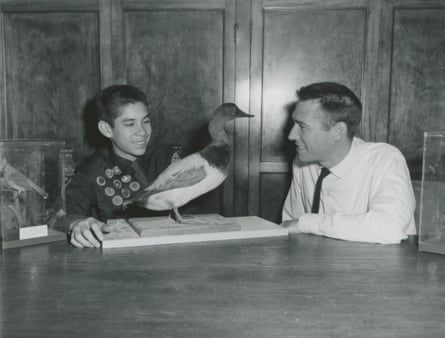 Black-and-white image of man in white shirt and thin black tie sitting at table with taxidermized bird, and boy wearing sash sewn with badges.