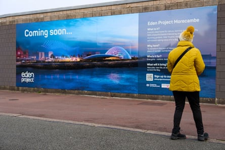 A woman in a warm yellow coat and woolly hat stops to look at a hoarding showing the planned Eden Project, with the words ‘Coming soon’ at the top