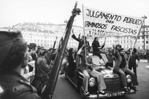 Young people ride on a car through Lisbon holding a sign saying 'Public trial of the fascist criminals' as soldiers line the streets