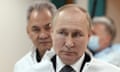 Putin shown from the shoulders up, looking to his right. Sergei Shoigu is pictured just behind him looking straight to the camera. Both appear to be wearing white protective coats over their clothes.