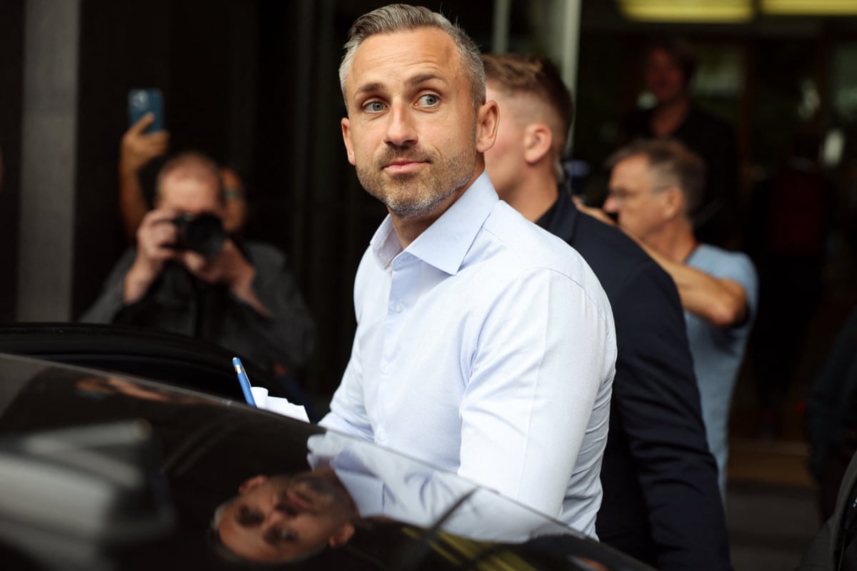 Ulrich Siegmund looks up as he leaves a press conference. He wears an open necked white shirt and stands behind a car as if he is about to get in.