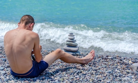 A man in swimming trunks sitting on a pebble beach photographing stones