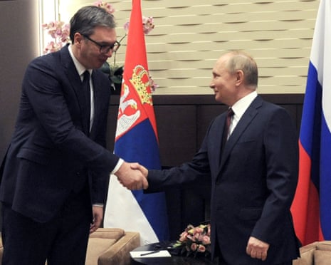 Putin and Vucic shake hands and smile, with Russian and Serbian flags behind them