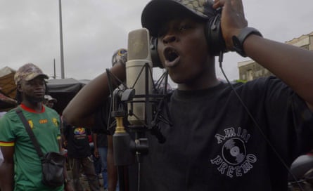 Young African musician, KMB, sings into microphone while wearing headphones, on the street.