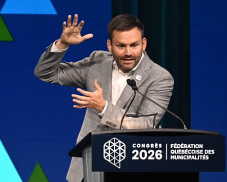 A man in a grey suit gestures with raised hand while speaking at a podium at the Federation Quebecois des Municipalities.