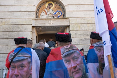 Men wearing Serbian flags as capes, each with a large picture of Ratko Mladic on them which appears across their backs, stand amongst people gathered outside a pale stone church which has a gold-decorated icon over the doorway.