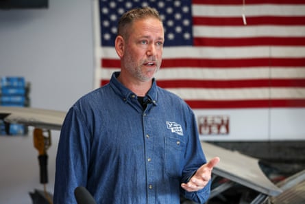 man in denim collared shirt speaks with hand gestured, an american flag hanging behind him