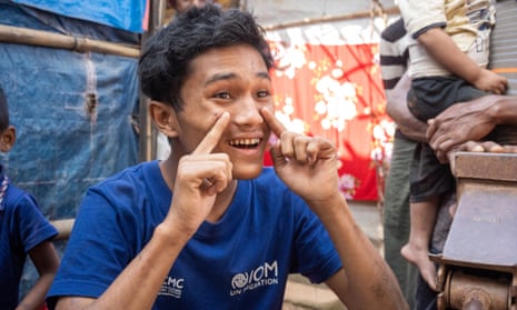 A smiling teenage boy gestures at his face. Shacks of bamboo and plastic sheeting can be seen behind him