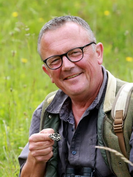 A man in a field smiling at the camera with a white butterfly with black spotted wings that has landed on his hand