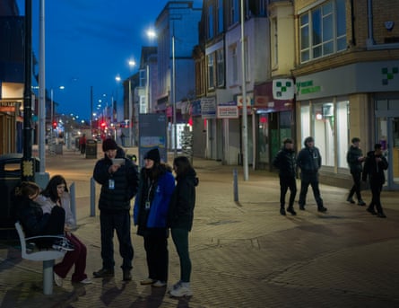 Two adult talking to three girls by a bench in a pedestrianised shopping area as a group of boys pass behind them