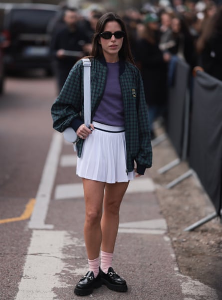 A young woman with long dark hair wearing sunglasses, a white tennis skirt, a navy T-shirt and a green check bomber jacket, with pink socks and chunky black loafers.