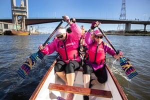 Caroline Mulcahy and Hazel Dean are paddling their dragon boat on the water in Melbourne's Docklands. They are holding brightly coloured paddles, and wearing bright pink life jackets. Their team is in the boat behind them and the Bolte Bridge is in the background