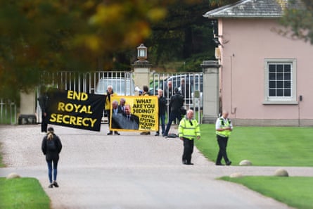 People hold banners saying ‘End royal secrecy’ and ‘What are you hiding? Royal Epstein inquiry now’ outside iron gates with two security officers in the foreground