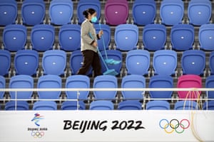 A maintenance worker wearing a face mask walks through an empty section of spectator stands near a logo for the Beijing 2022 Winter Olympics at a test event.