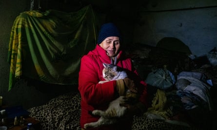 Angela in a red puffer coat and blue hat, sits on  her leopard-patterned blanket, cradling one of the cats she cares for. 