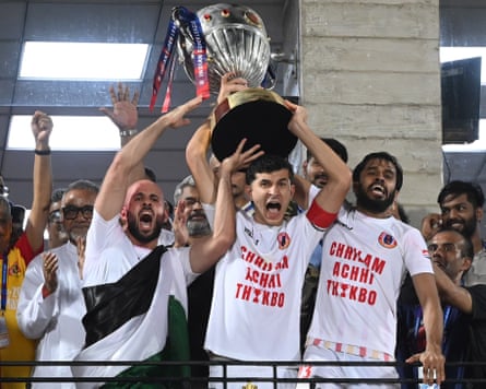 Players and staff of East Bengal FC stand on a balcony celebrate winning the Indian Super League, lifting the large silver trophy and punching the air.