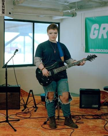 Girl in torn jeans holds an electric guitar with amps and mics behind her