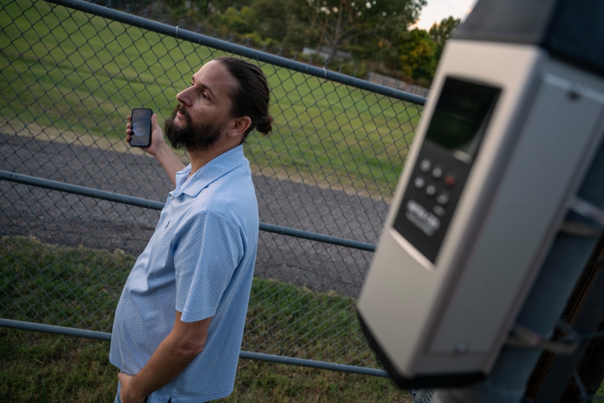 A man stands near a chain-link fence with a device mounted in the foreground.