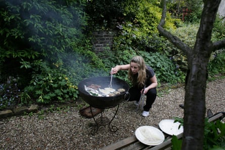 a young woman with long blond hair leans over a portable barbecue on a gravel path in a leafy, shady garden