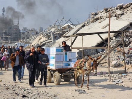 A donkey pulls a cart of goods as people walk along a path next to destroyed buildings.