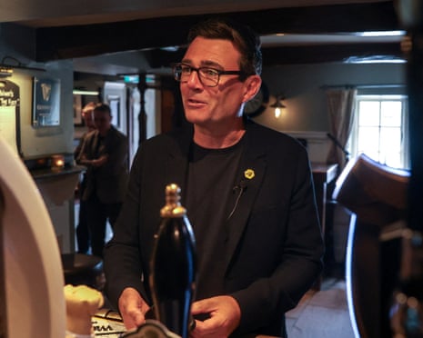 Andy Burnham in glasses and a black suit stands behind a bar with beer taps in a pub