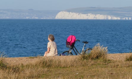 A woman sits next to a bike on the sand looking out to sea with white cliffs in the background