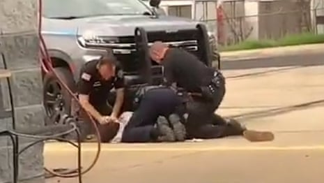 A grainy still from a video shows three officers on the ground next to a truck. Beneath them, barely visible, is a man they are arresting. The officer on the left has both hands on the man's head, holding it to the ground.