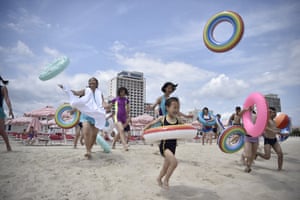 People in bathing costumes carry or throw rubber rings as they hasten across the sand towards the sea in front of a backdrop of red and white striped parasols and white highrise buildings