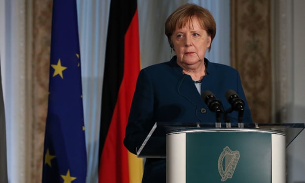 Angela Merkel standing at a microphone with flags in the background