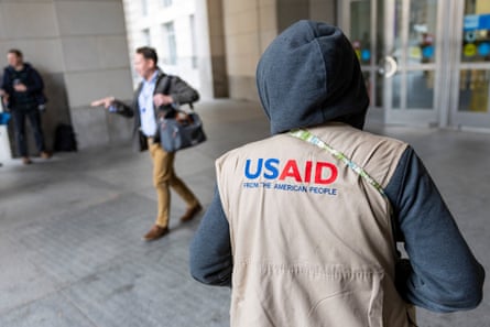 a person wears a vest that reads ‘USAID from the American people’ at a building entrance